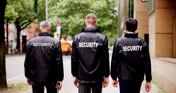 Security Guard In Front Of The Door Entrance. Young Security Guard Standing In Front Of The Glass Door Entrance